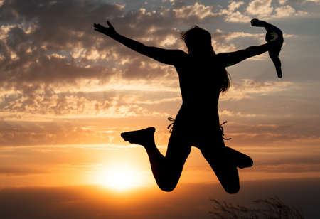 Young girl jumping silhouette with shawl on background of beautiful cloudy sky with orange sunset and rays of lightの写真素材