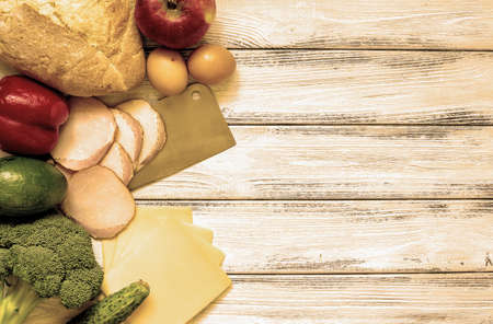 Set of products for cooking - apple, bell pepper, cucumber, fresh bread, eggs, sliced ham, broccoli, cheese, avocado and cleaver on white vintage wooden work surface warm filteredの写真素材