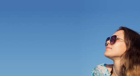Young pretty woman in sunglasses on blue sky background. Summer vacation concept with empty space for banner. Relax sun therapy copy space advertising. Beautiful tan brunette girl isolated on cler skyの写真素材