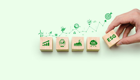 Wooden blocks displaying sustainability and ESG icons with a hand placing the ESG symbol, representing corporate responsibility, environmental impact, green growth, and sustainable strategyの写真素材