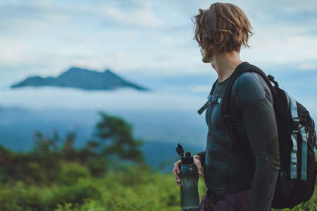 Outdoor portrait of Young man looking on Batur volcano and Agung mountain view at morning from Kintamani, Bali, Indonesia and drink waterの写真素材