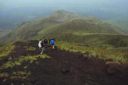 3 Men walk along the hill with backpacks and with white clouds and peak of volcano on backgroundの写真素材