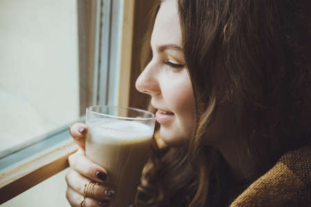 Beautiful young woman in casual wear dreaming with cup of hot coffee or tea over windowの写真素材