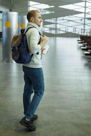 Passenger with Coffee Cup in Hand and Backpack  Sitting at the Hall of Airport Travel Concept