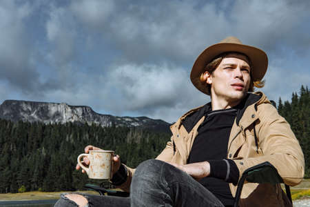 Attractive man in the hat is drinking coffee and enjoying the view of the lakeのeditorial素材