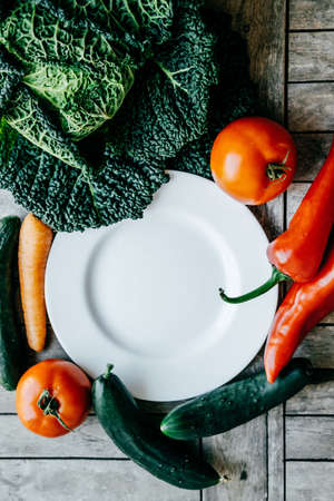 Fresh vegetables: kale, Carrot, pepper, tomato, cucumber around  empty plate on a wooden background....