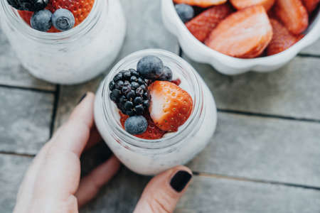 Top view of Woman hands with Pudding with chia seeds, yogurt and fresh fruits: Strawberries, blueberries and blackberries in glass jars on wooden backgroundの写真素材