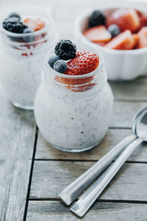 Vertical photo of Pudding with chia seeds, yogurt and fresh fruits: Strawberries, blueberries and blackberries in glass jars on wooden backgroundの写真素材