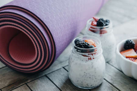 Sport and healthy concept. Pink yoga mat and Pudding with chia seeds, yogurt and fresh fruits: Strawberries, blueberries and blackberries in glass jars on wooden backgroundの写真素材