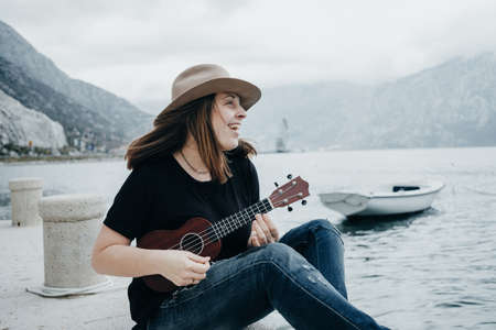 Young woman in a hat plays on a ukulele on the background of a bay in Montenegroの写真素材