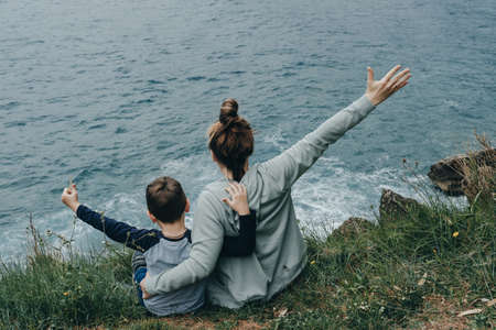 Back view of woman and her son raised their hands upwards sitting on the mountain above the sea. Freedom conceptの写真素材