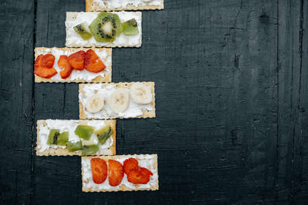 Snacks on a handmade Crackers with cheese and fruits: bananas, strawberries and kiwi on a black backgroundの写真素材