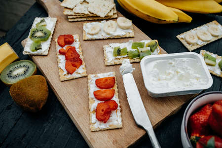 Light snack of water cracker sandwiches with cream cheese and fruits: bananas, strawberries and kiwi on a black background, Top viewの写真素材