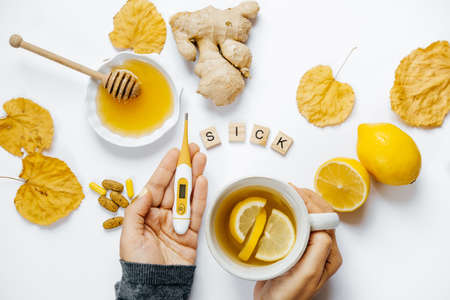 Female hands with tea and thermometer, lemon, ginger, honey, tablets and dry leaves on a white...