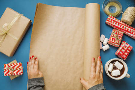 Woman hands unwrap the wrapping paper with space for text, hot cocoa with marshmallows, gifts, scissors on a blue table. top viewの写真素材