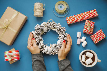 Woman hands holding wreath, hot cocoa with marshmallows, gifts, scissors on a blue table. top viewの写真素材