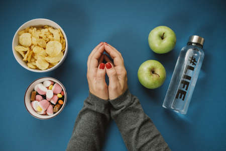 Top view with woman hands choosing apple or water against  candy, chips on blue background.の写真素材