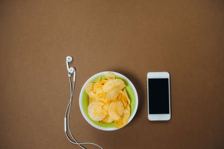 Top view of bowl with chips,phone and headphones on brown background. Unhealthy concept. の写真素材