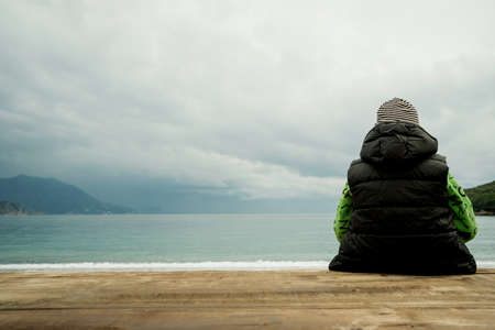 Back view of a boy sitting on a wooden board and looking at the seaの写真素材
