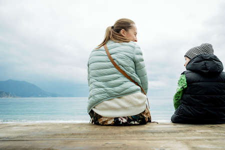 Back view of a boy and mom sitting on a wooden board and looking at the seaの写真素材