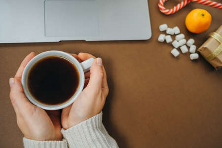 Women's hands with Coffee and marshmallows on Christmas background with Laptop and gifts on brown table. Top view, flat layの写真素材