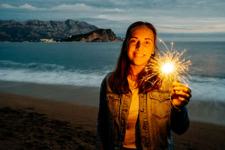 Outdoor photo of young happy smiling woman holding sparkler near the seaの写真素材