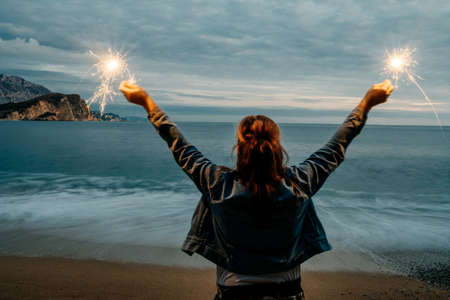 Back view of young happy smiling woman holding sparkler near the seaの写真素材