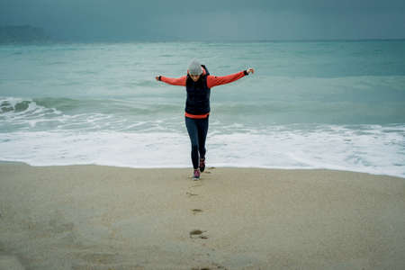 Joyful young woman standing on a cold winter's beach and stretching her armsの写真素材