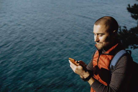 Man traveler in a red waistcoat watching world map on mobile phone while relaxing near sea の写真素材