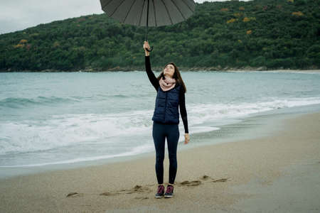 Joyful woman  breathing fresh air and standing with an umbrella in front sea in winter or autumnの写真素材