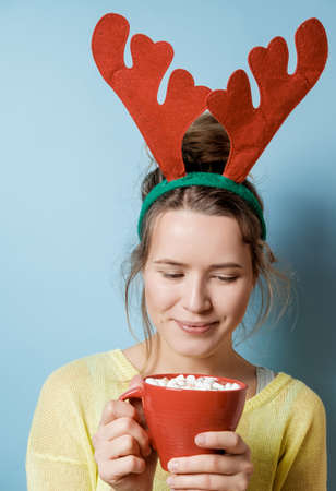 Beautiful young woman in Christmas horns with a red mug on a blue background. Studio shotの写真素材