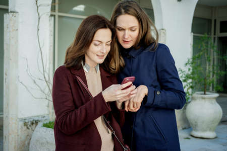 Two pretty young women in a blue and claret looking at phone in the city.
