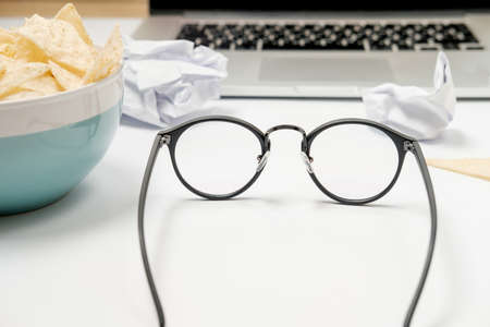 Workspace with spectacles and laptop, crumpled paper, stationery and chips on white table. Bad habits conceptの写真素材