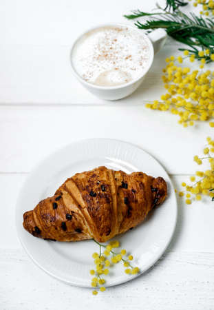 Chocolate croissant and cappuccino, decorated with mimosa flowers on a white table. Spring, Mother's Day concept.の写真素材