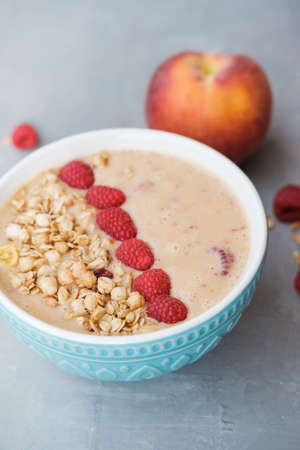 Peach smoothie bowl with granola and raspberry in a blue bowl on grey background. Top view. Healthy and colorful breakfastの写真素材