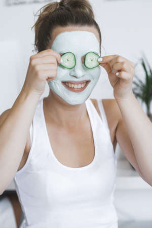 Young Pretty Blonde woman with facial mask posing with cucumber at home. Wellbeing concept, Morning skin care routine.の写真素材