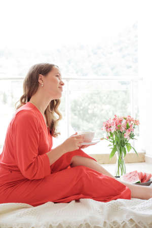 Young Blonde woman in a red robe  drinks coffee against wide window background and having lazy morningの写真素材