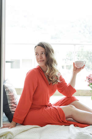 Young Blonde woman in a red robe  drinks coffee against wide window background and having lazy morningの写真素材