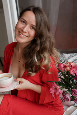 Portrait of Young Blonde woman in a red robe  drinks coffee against wide window background and having lazy morningの写真素材