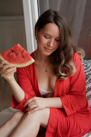Young Blonde woman in a red robe  eating a watermelon for breakfast  against wide window backgroundの写真素材