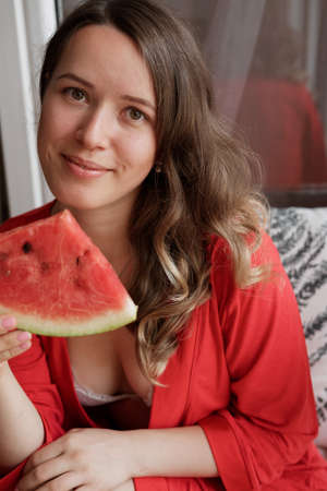 Young Blonde woman in a red robe  eating a watermelon for breakfast  against wide window backgroundの写真素材