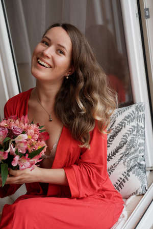 Young Blonde woman in a red robe with flowers against wide window background having lazy morningの写真素材
