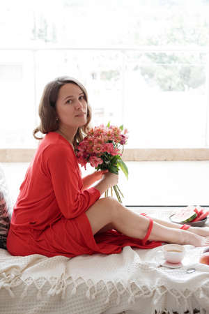 Young Blonde woman in a red robe with flowers against wide window background having lazy morningの写真素材