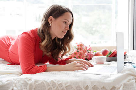 Pretty blonde woman in a red robe working on laptop on the bed and drinks coffee against window backgroundの写真素材