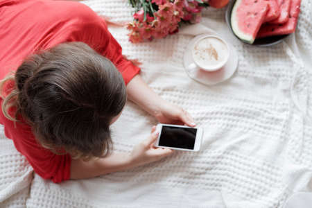 Top view of a Pretty blonde woman in a red robe using phone on the bed and drinks coffee against window backgroundの写真素材