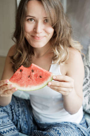 Young Blonde woman in blue pajamas on the bed  eating a watermelon for breakfast  against window backgroundの写真素材