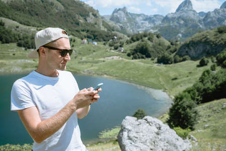 Traveler man in a cap and sunglasses standing using smartphone and see beautiful scenery   mountain landscape in Montenegroの写真素材