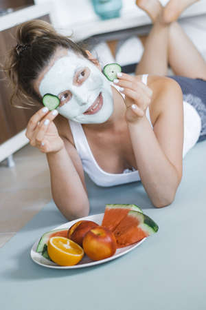 Young Pretty Blonde woman with towel on head and facial mask with cucumber eating watermelon at home. Wellbeing concept, Morning skin care routine.の写真素材