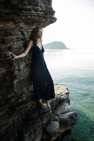 Young curly blonde woman in long dress against rock by the sea. Slow motionの写真素材