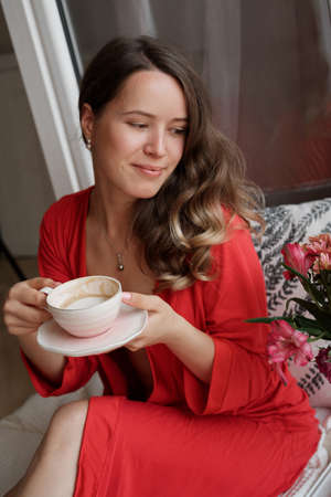 Portrait of Young Blonde woman in a red robe  drinks coffee against wide window background and having lazy morningの写真素材